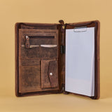 An open brown leather portfolio folder or notebook cover on a warm beige background, showing interior pockets, a knife sheath, and a lined notepad with a clip.