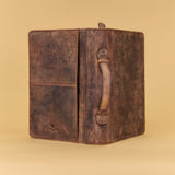 A distressed brown leather journal with a single leather handle and a front pocket, shown open on a solid beige background.