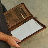 A person holds open a brown leather notebook cover or organizer, showing lined paper inside alongside tools such as a small knife, hook, and pen, against a textured gray wall.