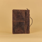 A dark brown distressed leather journal with a front pocket and a black strap closure, standing upright against a solid beige background.