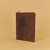 A dark brown distressed leather zippered notebook or journal standing upright on a warm beige studio background.
