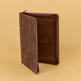 A dark brown leather journal or notebook cover standing open at a slight angle, showing its interior lining, set against a solid warm beige background.