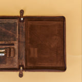 An open brown leather travel wallet or document holder with multiple card slots, a zippered pocket, and a blank interior page, shown on a warm beige background.