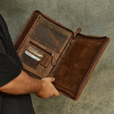 A person in a black shirt holds open a brown leather portfolio or document case, showing interior card slots and pockets against a textured gray background.