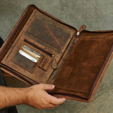 An open brown leather portfolio folder or document case held by a person's hand, showing interior card slots, pockets, and a zipper compartment against a textured gray background.