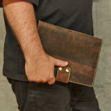 A person in a black t-shirt holding a brown distressed leather notebook cover or document holder with brass snap buttons, set against a textured gray wall.