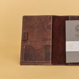 A brown leather journal cover shown open on a warm beige background, holding a gray journal with a white paper band and a small leather strap closure.