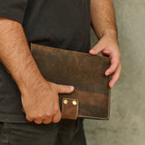 A person holding a dark brown distressed leather tablet sleeve or folio with brass snap buttons, shown from the chest down against a textured gray wall.