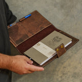 A person holding an open brown leather notebook cover or organizer, which contains a gray notebook and a pen, against a textured gray concrete background.