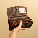 A hand holds an open Leather Ladies Wallet Alice by Vintage Leather Sydney, a womens vintage waxed brown leather wallet with multiple compartments, card slots, a trifold design, and a clear ID window. The background is plain beige.