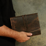 A person in a black shirt holds a dark brown distressed leather notebook or journal cover with an elastic strap closure against a textured gray background.