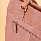 Close-up of a brown leather bag showing a front zip pocket with a pull tab and brass-tone hardware against a warm beige background.