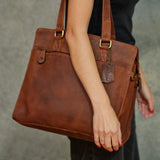 A person holding a large brown leather shoulder bag with gold-tone hardware and a hanging brand tag. The person is wearing a black top and dark pants, with red-painted fingernails visible against a gray textured background.