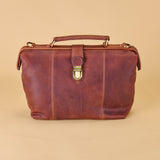 A brown distressed leather briefcase-style handbag with a top handle, front strap closure, and brass-tone hardware, photographed on a plain warm beige background.