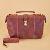 A distressed brown leather briefcase-style handbag with a top handle, front strap closure, and a matching wristlet pouch placed in front, photographed on a plain warm beige studio background.