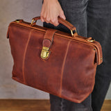 A person holding a brown leather briefcase with gold-tone hardware, shown from the side against a neutral indoor background.
