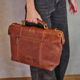 A person in dark gray jeans holds a brown distressed leather briefcase-style bag with gold-tone hardware against a neutral gray wall.