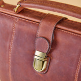 Close-up of a brown leather briefcase or satchel with a brass buckle closure and top handle, shown against a warm beige background.