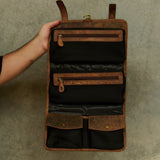 A brown leather and black mesh hanging organizer pouch with multiple zippered compartments, held open by a person's hand against a textured gray wall.