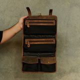 A brown and black leather and canvas roll-up organizer pouch held open by a person's hand against a textured gray concrete wall.