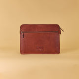 A brown leather laptop sleeve with a front zippered pocket and a small embossed logo patch, set against a solid beige background.
