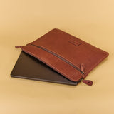 A brown leather laptop sleeve with a zipper closure, shown partially open with a dark laptop inside, set against a solid beige background.