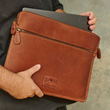 A person holding a brown leather laptop sleeve with a laptop partially inserted, shown against a neutral gray textured background.