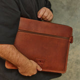 A person in a black t-shirt holding a brown leather laptop sleeve or document bag with a top zipper and a small logo patch on the front, set against a textured gray concrete wall.