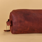 A brown textured leather cosmetic pouch with a zipper closure and hanging leather pulls, photographed on a solid beige background.