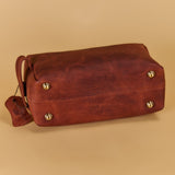 A reddish-brown leather shoulder bag with brass-tone metal studs and a matching leather tag, photographed on a solid beige background.