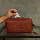 A brown leather travel pouch with a hand inserting a white bottle into its top zipper, set on a glass table against a textured gray wall.