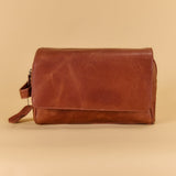 A brown leather clutch or pouch with a fold-over flap and a small wrist strap, photographed against a solid beige background.