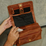 A person's hands open a brown leather travel organizer pouch, showing its black interior lining, mesh pocket, and gold-tone hardware against a gray concrete wall.