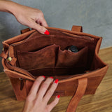 A brown leather tote bag shown open with a hand inside, revealing interior organizer pockets. Another hand with red nail polish is visible at the bottom, and the bag sits on a wooden surface against a gray wall.