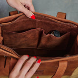 Close-up of a brown leather tote bag being opened by two hands with red nail polish, showing the interior organization with multiple pockets and a small bottle in a side pocket.