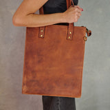 A person holding a large brown leather tote bag with gold-tone hardware against a textured gray background.