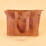 A brown leather tote bag with dual shoulder straps, a front slip pocket, and a small hanging leather tag, photographed against a solid beige background.