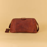 A brown distressed leather crossbody bag with a top strap closure and a small hanging leather tag, set against a solid beige background.