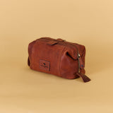 A brown leather travel pouch with a top zipper and a small brand patch on the front, set against a solid beige background.