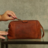 A brown leather travel pouch with a top zipper and strap, held by hands over a glass table against a textured gray wall.