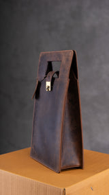 The Leather Wine Carry Bag Sudbury by Vintage Leather Sydney, a brown leather tote with a rectangular cut-out handle and gold clasp, stands upright on a cardboard box against a dark gray textured background.