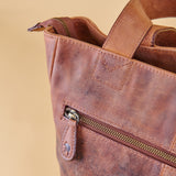 Close-up of a distressed brown leather tote bag with a front zip pocket and a top zipper, set against a solid beige background.