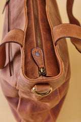 Close-up of a brown leather handbag showing the zipper closure, leather pull tab with a small blue logo, and brass-tone hardware against a warm beige background.