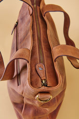 A close-up of a brown leather shoulder bag showing the interior zipper, strap attachments, and a brass ring detail against a warm beige background.