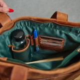 Close-up of the inside of a brown leather bag, showing a green satin-lined compartment with a bottle holder, card slots, a blue pen, and a pen loop. A hand with red painted nails is visible holding the bag open.