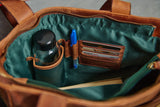 Close-up of the interior of a brown leather bag with a dark teal satin lining. The bag is shown open, containing a bottle in a side pocket, a blue pen in a pen loop, cards in a card holder, and a notebook.
