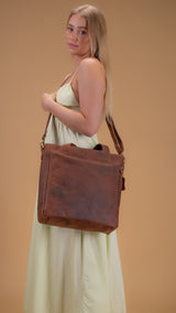 A woman in a light yellow dress stands against a tan background, holding the Vintage Leather Sydney Leather Work Tote Bag Parsley. She looks toward the camera with a relaxed expression.