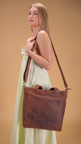 A woman in a light green dress stands sideways, holding the Leather Work Tote Bag Parsley by Vintage Leather Sydney, featuring short handles and a long strap, against a solid beige background.
