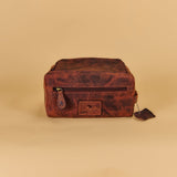 A distressed brown leather zippered pouch or small bag with a front patch logo, photographed on a plain beige studio background.