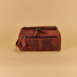 A brown distressed leather toiletry bag with a top zipper and a front zippered pocket, set against a solid beige background.